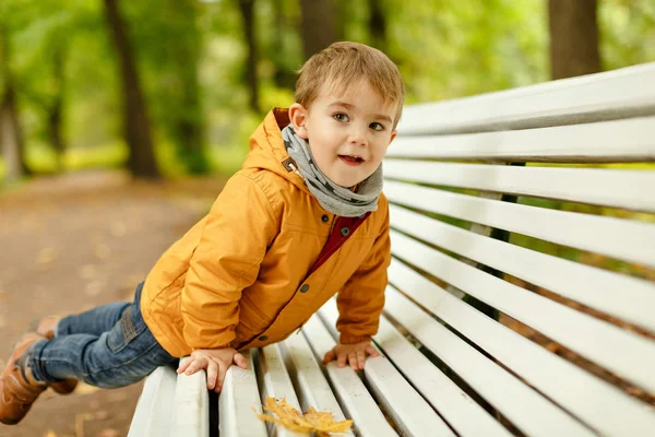 Pequeño niño adorable con chaqueta amarilla acostado en el banco, y rej — Foto de Stock