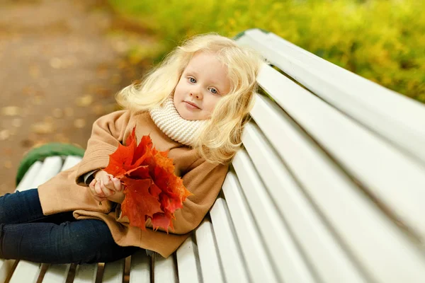 Roztomilý nadýchané blond holčička v kabátě seděl na lavičce v t — Stock fotografie