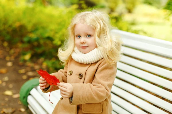 Kleine süße flauschige blonde Mädchen in einem Mantel sitzt auf einer Bank in t — Stockfoto