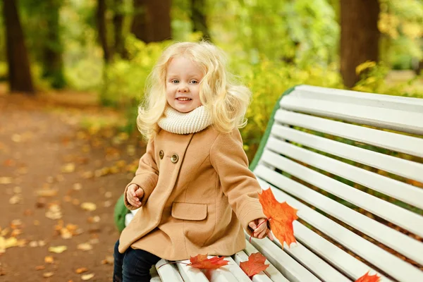 Little cute fluffy blonde girl in a coat sitting on a bench in t — Stock Photo, Image