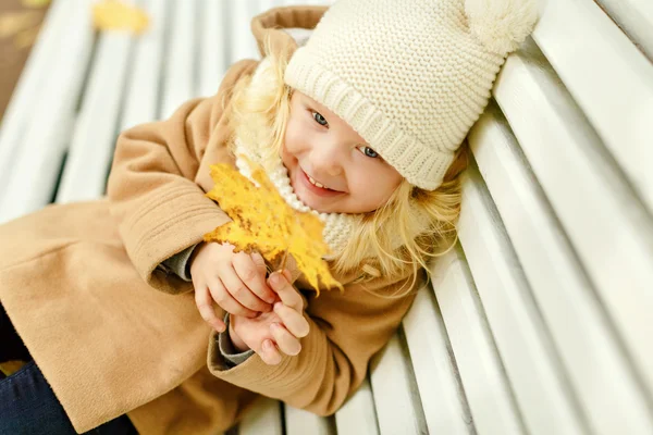 En liten pen jente i en beige frakk sitter på en benk og ler. – stockfoto