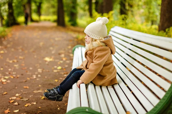 Fetiță drăguță într-o haină bej așezat pe o bancă și trist la — Fotografie, imagine de stoc
