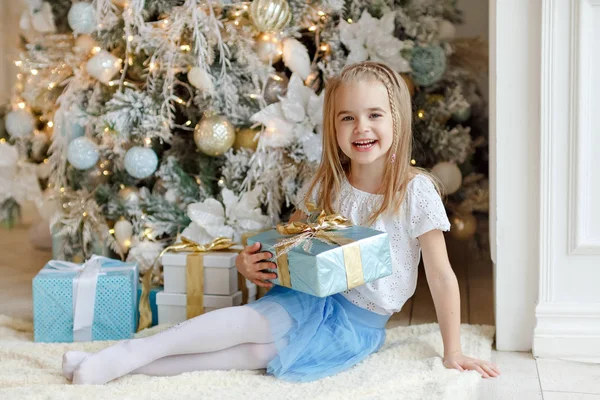 Hermosa niña sentada cerca de un árbol de Navidad y sonríe i — Foto de Stock