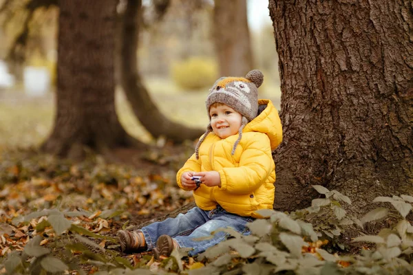 Bambino in giacca gialla sorride in autunno, seduto sotto — Foto Stock