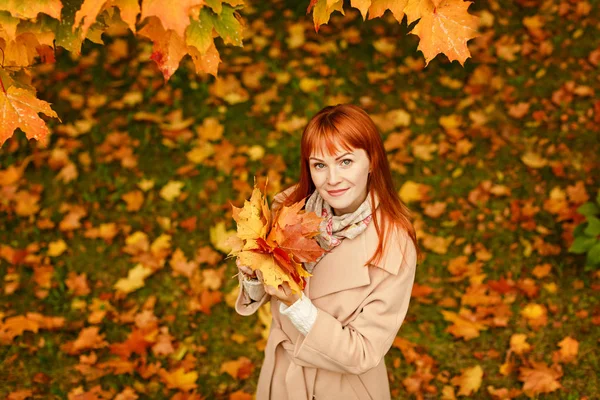 The red-haired girl in a beige coat smiles on autumn background — Stock Photo, Image