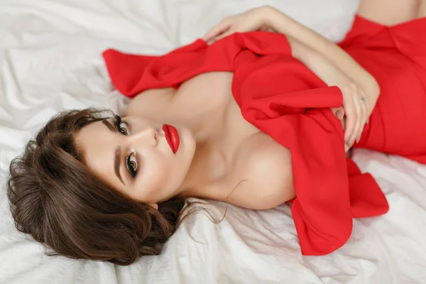 Elégante fille brune sexy dans une robe rouge se trouve sur un lit — Photo