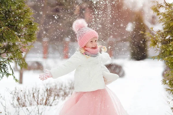 Little girl in a pink skirt in winter smiles in the park and thr — Stock Photo, Image