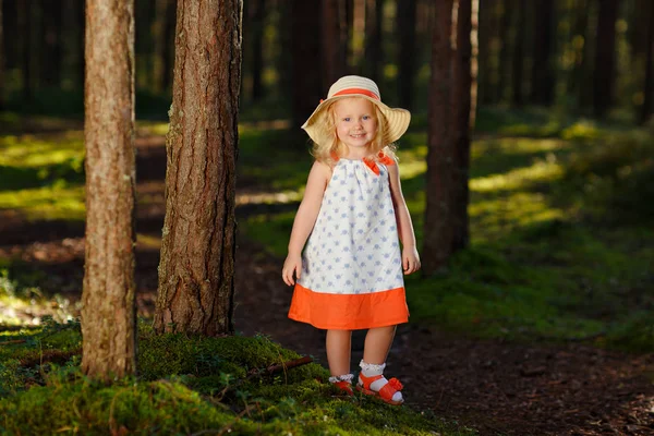Ein kleines blondes Mädchen in einem hellen Kleid und Hut geht in einer Kiefer f — Stockfoto