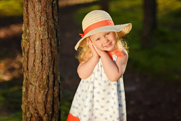 A little blonde girl in a bright dress and hat walks in a pine f — Stock Photo, Image