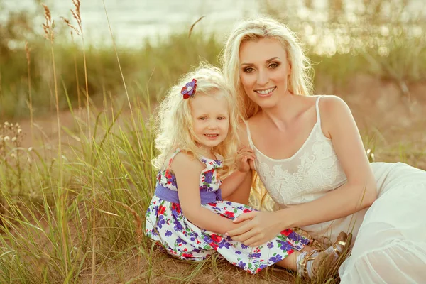 Ein kleines blondes Mädchen und ihre Mutter lächeln auf Sand und Gras — Stockfoto