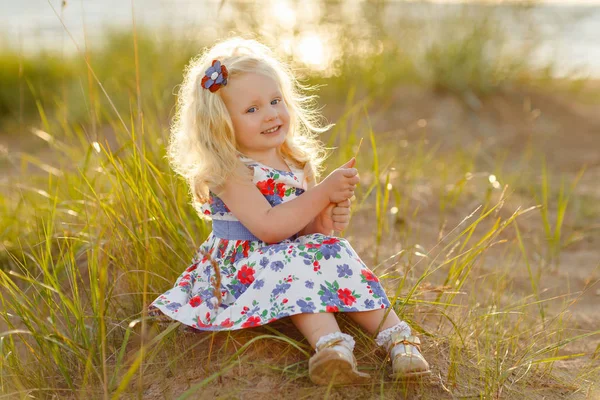 Pequeña chica rubia rizada se sienta y sonríe en la arena y la hierba en su —  Fotos de Stock