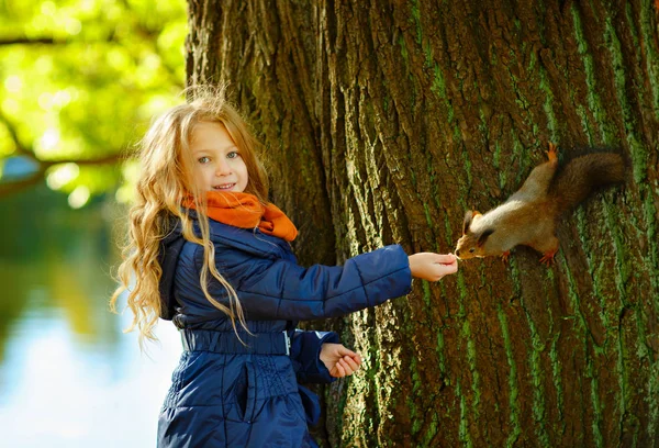 Uma linda garota loira de 6 anos sorrindo e alimentando um esquilo na floresta de outono — Fotografia de Stock