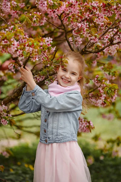 A cute blonde girl smiles against a background of rose sakura bu — Stock Photo, Image