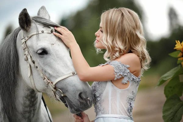 Menina loira bonita no vestido acaricia um cavalo cinza na natureza em — Fotografia de Stock