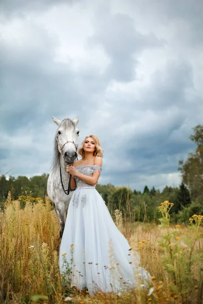 Linda menina loira esbelta no vestido abraçando um cavalo cinza, para fora — Fotografia de Stock