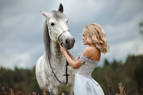 Beautiful slender blonde girl in dress looks at gray horse, on n — Stock Photo, Image