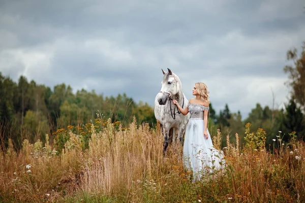 Όμορφη λεπτή ξανθιά κοπέλα στο φόρεμα που αγκαλιάζει ένα γκρίζο άλογο, έξω — Φωτογραφία Αρχείου