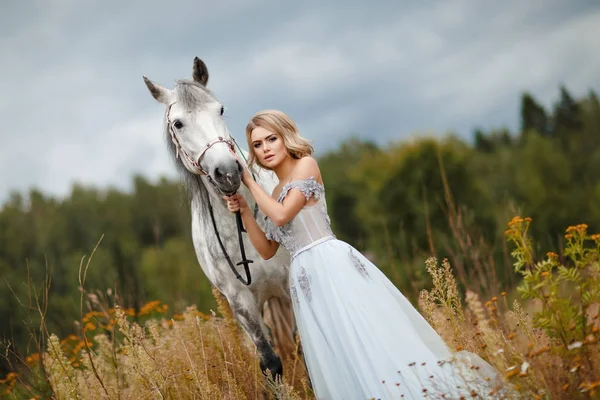 Linda menina loira esbelta no vestido abraçando um cavalo cinza, outd — Fotografia de Stock