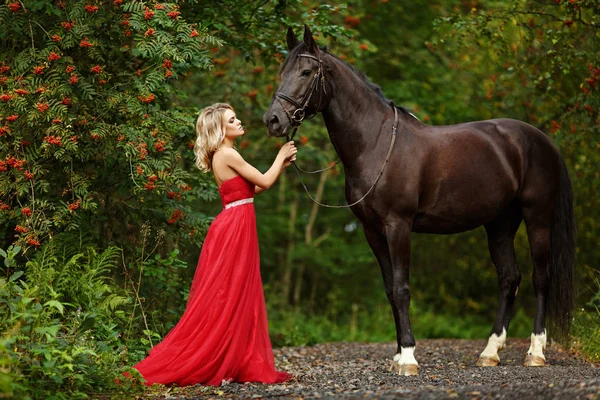 Belle fille blonde élancée en robe rouge embrassant un cheval noir — Photo