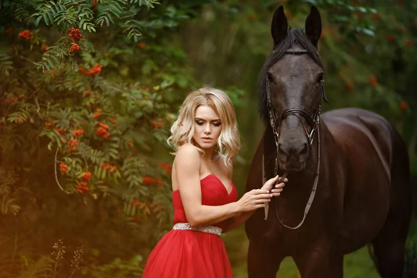 Beautiful slender blonde girl in red dress hugging a black horse — Stock Photo, Image