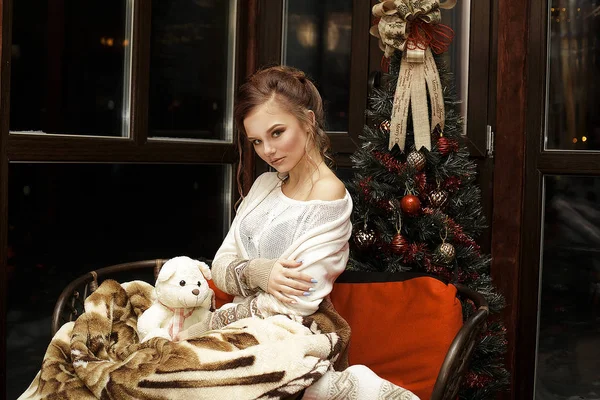 Красивая блондинка обнимается сидя расслабляясь с теплым одеялом на диване рядом с елкой и темным окном — стоковое фото