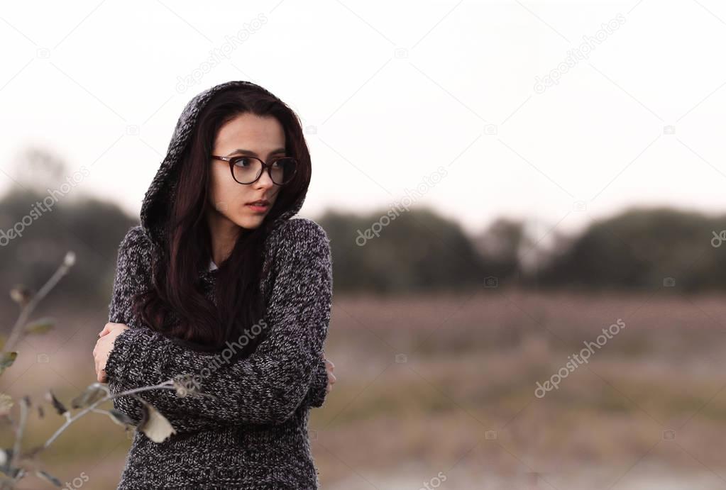 Lonely alone beautiful girl in warm sweater with hood looking aside in autumn field background. Picture of pretty depressed sad unhappy girl with glasses. Alone girl having drug addiction.