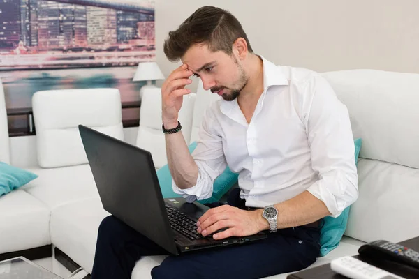 Thinking handsome man working t laptop at home. Young man relaxing on the sofa with a laptop. Multitasking.Handsome young man wearing glasses and working with touchpad while sitting on couch in office