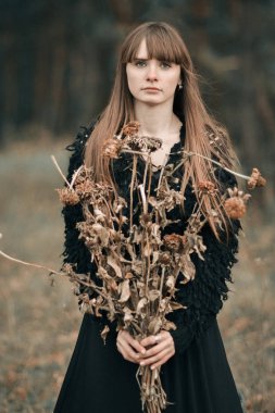 Kuru sonbahar çiçekler buket tutarak uzun saçlı güzel kız üzgün, mutsuz, ciddi. Koyu renkleri sonbahar karanlık ormanın içinde kuru çiçekler ile siyah elbiseli güzel kız resim.