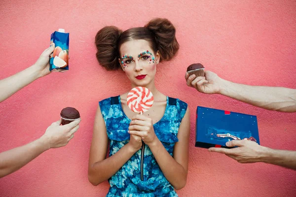 Hermosa chica de moda con dos bollos de pelo un maquillaje creativo con una enorme piruleta dulce rosa en las manos. Muchas manos sobre fondo rosa alrededor de una chica sosteniendo pasteles, jugo, caramelos, dulces sobre fondo — Foto de Stock