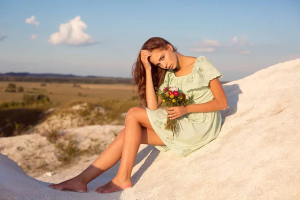 Όμορφο κορίτσι που κάθεται σε λευκό λόφους στο ηλιοβασίλεμα. Λίγο ηλιοκαμένο κορίτσι με μπουκέτο από λουλούδια που κάθεται στο βουνό με καταπληκτική θέα. Πανέμορφο κορίτσι σε πράσινο φόρεμα καλοκαίρι στη φύση — Φωτογραφία Αρχείου