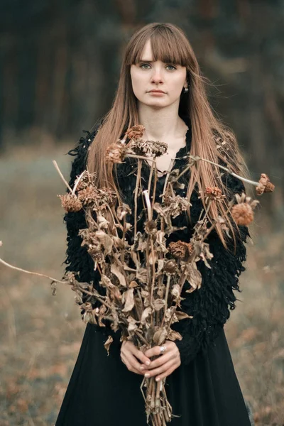 Chica hermosa triste, infeliz, seria con el pelo largo sosteniendo un ramo de flores secas de otoño. Imagen en colores oscuros de chica bonita en vestido negro con flores secas en el bosque oscuro de otoño . — Foto de Stock
