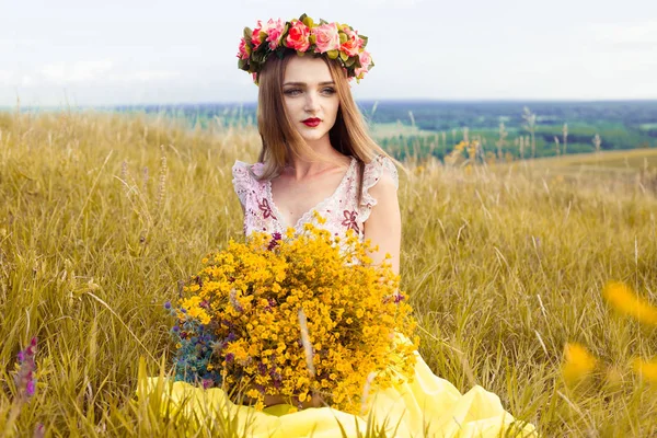 Hermosa chica bonita de moda en vestido en el campo de flores. Linda chica con corona de flores en la cabeza y ramo de muchas flores amarillas sentadas en el campo amarillo prado en verano . — Foto de Stock