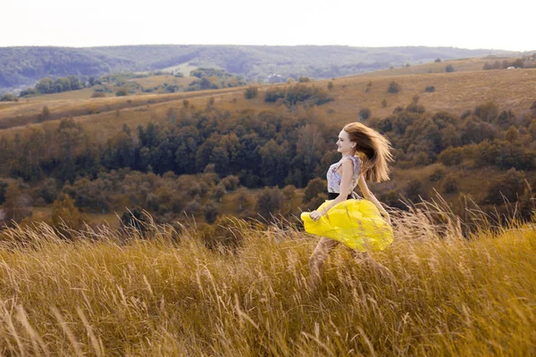Feliz juguetona niña bonita corriendo en el campo con verde, trigo amarillo en el camino a la buena vida.Feliz aventura en verano, verano.Chica sonriente alegre en vestido amarillo claro corriendo en el campo — Foto de Stock