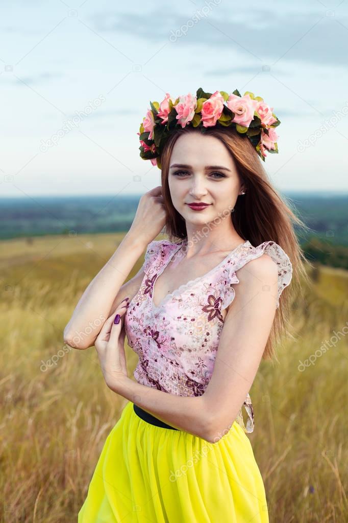 Beautiful fashionable pretty gorgeous girl in dress on the flowers field. Nice girl with wreath of flowers on her head and bouquet of many yellow flowers sitting on meadow yellow field in summertime.