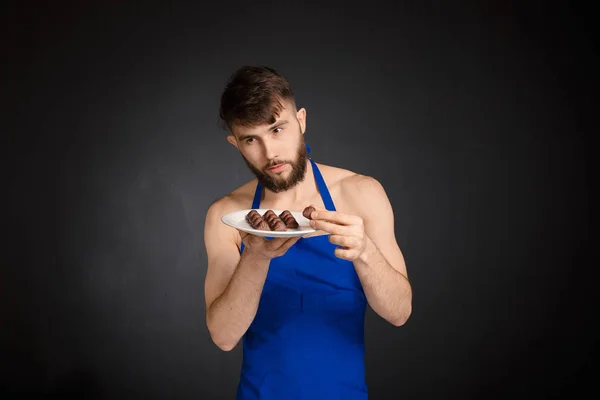 Hete sexy naakte knappe man met chocolade, chocolade snoepjes. Glimlachend naakte knappe man dragen donker blauw schort Houd een witte plaat met veel snoep, snoepjes chocolade. Knappe banketbakker. — Stockfoto