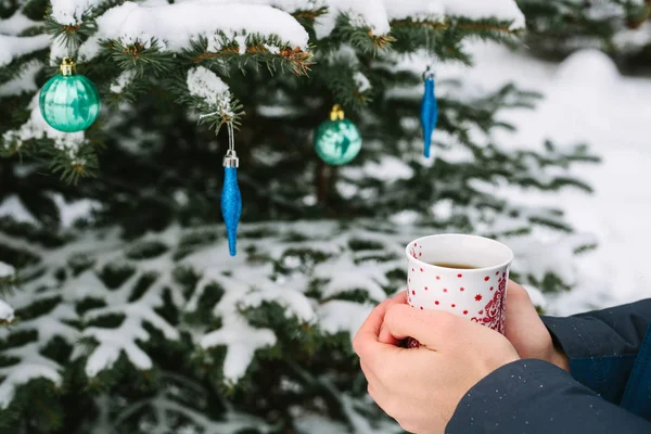 Man holding a cup of hot tea in his hands next to the Christmas tree in winter forest in the New Year holidays — Stock Photo, Image