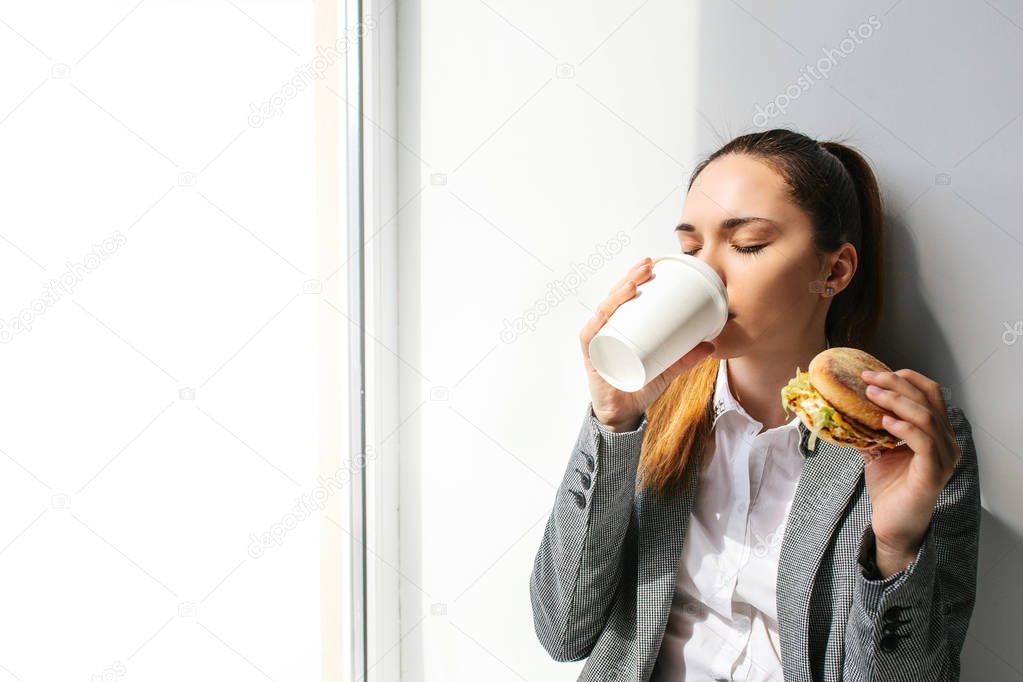 A young beautiful girl drinks coffee next to a window and eats a burger in a break between work. Fast food.