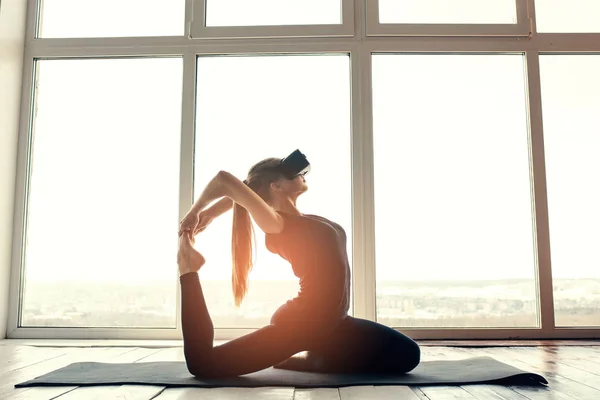 Una joven hermosa chica en gafas de realidad virtual hace yoga y aeróbic de forma remota. Concepto de tecnología futura. Tecnología de imagen moderna. Clases en deportes individuales remotamente . —  Fotos de Stock