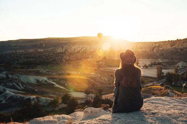 A tourist girl in a hat sits on a mountain and looks at the sunrise and balloons in Cappadocia. Tourism, sightseeing, Turkey. — Stock Photo, Image