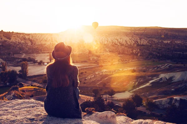 Een toeristische meisje in een hoed zit op een berg en kijkt naar de zonsopgang en ballonnen in Cappadocië. Toerisme, attracties, Turkije. — Stockfoto