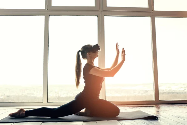 Una joven hermosa chica en gafas de realidad virtual hace yoga y aeróbic de forma remota. Concepto de tecnología futura. Tecnología de imagen moderna. Clases en deportes individuales remotamente . —  Fotos de Stock