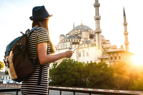 Una joven y hermosa viajera en un sombrero con una mochila está viendo un mapa junto a la mezquita azul, la famosa vista de Estambul. Viajes, turismo, turismo . — Foto de Stock