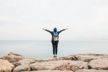 Denizin yanında bir sırt çantası ile şapkalı bir kız Gezgin ellerini yukarı yükseltir. Seyahat, dinlenme, hiking, özgürlük.