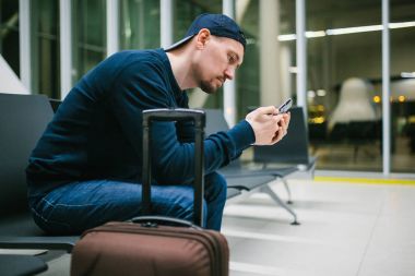 Genç bir adam ile bir bavul Havaalanı bekleme odasında oturur ve bir cep telefonu kullanır. Gece uçuşu, transfer, havaalanında bekliyor.