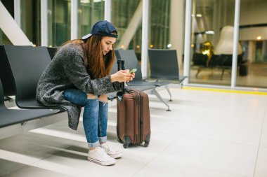 Bir çanta ile rahat tarzı bir genç güzel kız Havaalanı bekleme odasında oturur ve bir cep telefonu kullanır. Gece uçuşu, transfer, havaalanında bekliyor.