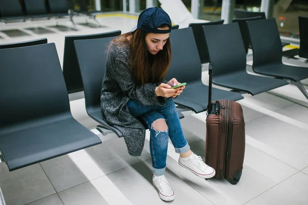 Bir çanta ile rahat tarzı bir genç güzel kız Havaalanı bekleme odasında oturur ve bir cep telefonu kullanır. Gece uçuşu, transfer, havaalanında bekliyor.