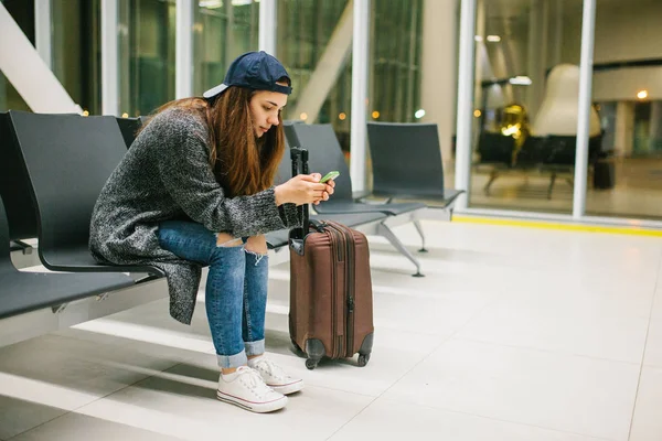 Bir çanta ile rahat tarzı bir genç güzel kız Havaalanı bekleme odasında oturur ve bir cep telefonu kullanır. Gece uçuşu, transfer, havaalanında bekliyor.