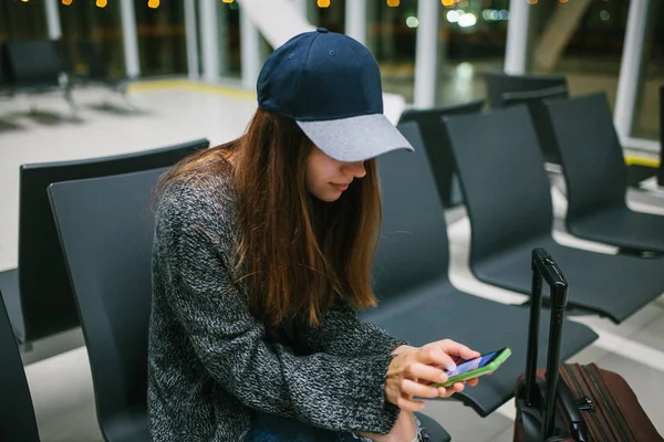 Bir çanta ile rahat tarzı bir genç güzel kız Havaalanı bekleme odasında oturur ve bir cep telefonu kullanır. Gece uçuşu, transfer, havaalanında bekliyor.