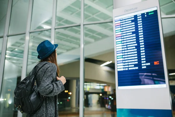 Bir sırt çantası içinde bir şapka ile genç kız Gezgin bilgi yönetim kurulu havaalanında bakar. Uçuş hakkında bilgi alma. Transfer.