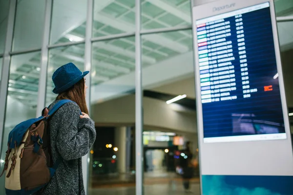 Bir sırt çantası içinde bir şapka ile genç kız Gezgin bilgi yönetim kurulu havaalanında bakar. Uçuş hakkında bilgi alma. Transfer.
